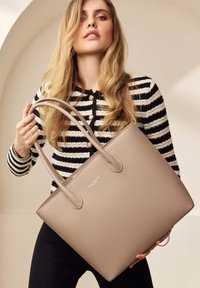 Sac fourre-tout en cuir beige avec deux poignées allongées et surface texturée. Présente un logo de marque discret à l'avant. Forme rectangulaire standard.
