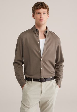 Bruine blouse met lange mouwen, gestructureerde stof en witte knopen, gedragen over een wit hemdje, gecombineerd met lichtbeige broek en een zwarte riem.