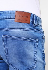 Jeans de denim azul con una etiqueta de cuero que dice "ONLY & SONS", con una textura suave y un diseño estándar de bolsillo trasero.