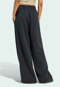 Osoba odjevena u široke crne hlače širokih nogavica i bež kratki top, stoji s rukama u džepovima okrenuta leđima.