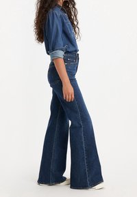 Femme en chemise en jean bleue et jean taille haute à jambes larges, debout de profil, manches de chemise retroussées et portant des baskets blanches.