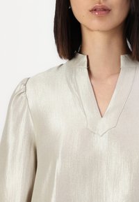 Blusa color oro chiaro con una leggera lucentezza, scollo a V e maniche a sbuffo con delicate cuciture. Texture liscia e un drappeggio elegante.