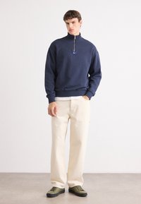 Sudadera con cremallera de color navy y cuello alto, combinada con pantalones de color crema y zapatillas verdes. Presenta un pequeño logo en el pecho.