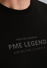 Crna pamučna majica s okruglim izrezom, koja ima natpis "NEOGRANIČENA SLOBODA PME LEGEND AMERICAN CLASSIC" u hrabrom, kontrastnom printu.