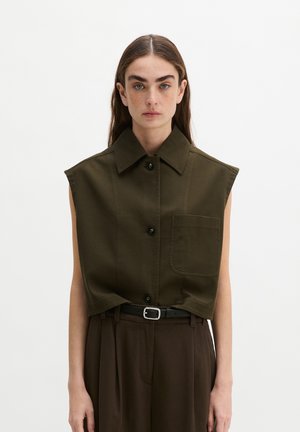 Donna con lunghi capelli castani che indossa una camicia verde oliva senza maniche con grandi tasche frontali e pantaloni marrone scuro con cintura nera.