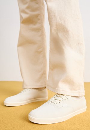 Person iført off-white afslappede sneakers med snørebånd og løstsiddende lyse beige bukser, stående på en sennepsgul gulv.