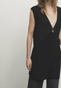 Vrouw in een mouwloze zwarte gebreide top met diepe V-hals en een zilveren hangersnoer, in combinatie met donkere broeken tegen een lichte achtergrond.