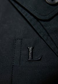 Zwarte stof met een gladde textuur, voorzien van een genaaide zak en een geborduurd letter "L" in een donkerdere tint. Eenvoudige knoopdetail zichtbaar.