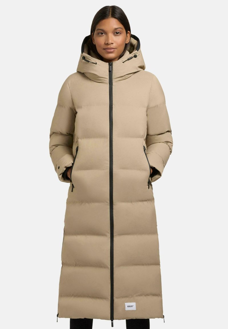 Cappotto puffer beige con silhouette lunga, dotato di cappuccio, zip nera e tasche laterali. Tessuto testurizzato, isolato e con cuciture a scatola.