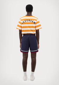 En orange og hvid stribet t-shirt med "ICECREAM" trykt på ryggen, parret med navy shorts med hvid og rød kant samt hvide sko.