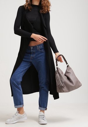 Kvinna som bär en svart lång cardigan, svart topp, blå uppvikta jeans, vita sneakers och håller en taupe läderhandväska med fransdetaljer.