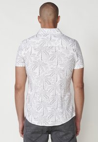 Camisa de manga corta en blanco con un patrón de palmeras grises. Cuenta con un cuello y un dobladillo curvado. Textura de tela suave.