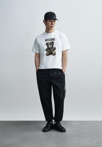 Άσπρο βαμβακερό t-shirt με γραφικό αρκούδας και το κείμενο "MOSCHINO", σε συνδυασμό με μαύρο παντελόνι με τσεπάκι με φερμουάρ και μαύρα αθλητικά παπούτσια.
