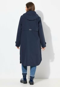 Manteau imperméable bleu marine avec capuche, manches longues et coupe ample. Comprend un détail de logo brodé dans le dos. Associé à un jean bleu et des bottes.
