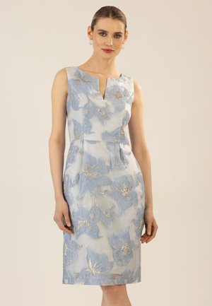 Frau in einem ärmellosen, knielangen Kleid mit blauer und goldener Blumenstickerei, stehend vor einem schlichten beigen Hintergrund.