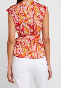 Blomstret bluse i pink med røde, orange og hvide bladmønstre. Materialet er let og med en tætsiddende talje og korte ærmer.