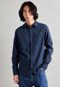 Camisa azul marino con rayas verticales en azul claro, diseño de botones, mangas largas, un bolsillo en el pecho y detalle de logo verde.