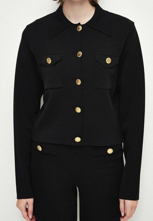 Veste noire à manches longues boutonnée et pantalon assorti avec des boutons décoratifs dorés portés par une personne, cadrée au niveau du cou et des hanches.