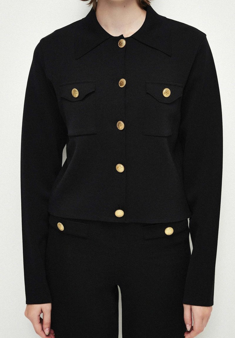 Veste noire à manches longues boutonnée et pantalon assorti avec des boutons décoratifs dorés portés par une personne, cadrée au niveau du cou et des hanches.