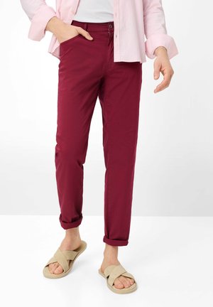 Person iført opsmøgede bordeauxbukser, lyserød skjorte og beige slip-on sandaler, stående med den ene hånd i lommen.