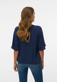 Donna con lunghi capelli castani ondulati, che indossa una blusa blu navy a maniche corte e jeans blu, rivolta di spalle su uno sfondo bianco uniforme.