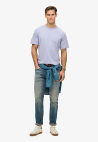 Camiseta de color púrpura claro con un pequeño gráfico, combinada con jeans de color azul claro remangados en los puños y zapatillas blancas. Chaqueta de mezclilla atada en la cintura.
