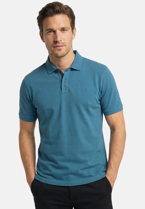 Man met kort bruin haar, gekleed in een blauwgroen poloshirt met korte mouwen en zwarte broek, staand met handen in de zakken tegen een witte achtergrond.