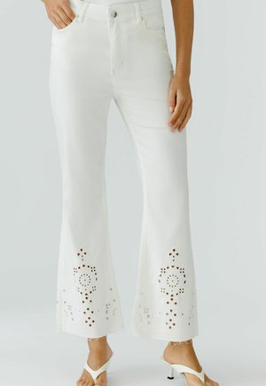 Femme portant un jean évasé blanc avec des motifs floraux découpés près des ourlets effilochés et des sandales blanches à talons.