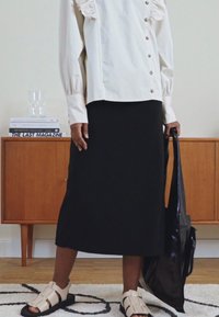 Camisa creme com botões e detalhes em babados, combinada com uma saia preta até ao joelho. Usando sandálias bege e segurando uma mala tote preta.