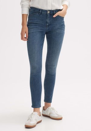 Skinny jeans in medium blue denim with a smooth texture, featuring a zip fly and side pockets. Paired with white sneakers and a buttoned shirt.