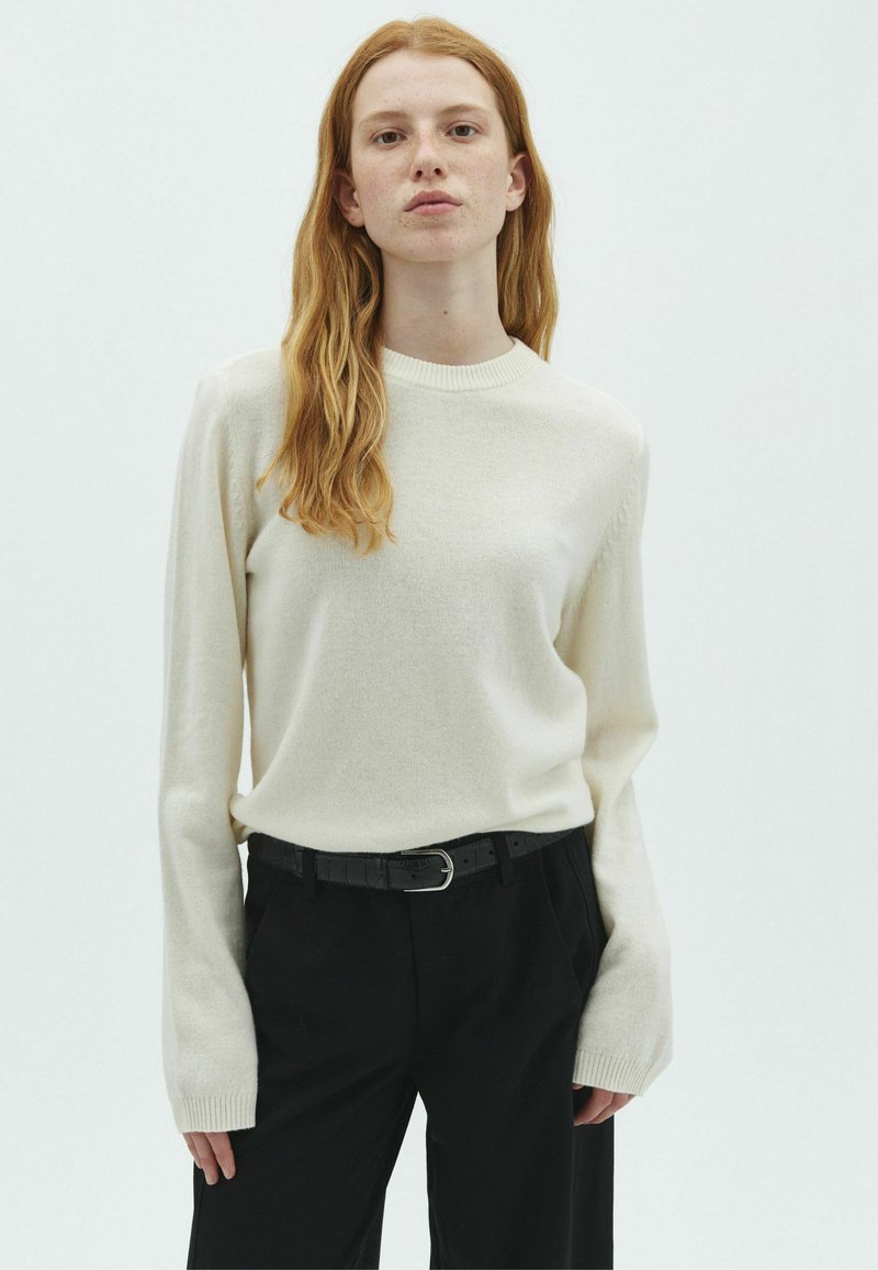 Jeune personne aux longs cheveux roux portant un pull blanc et un pantalon noir avec une ceinture noire, debout devant un fond blanc uni.