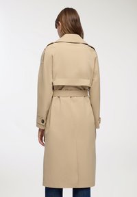 Beżowy płaszcz trench z paskiem w talii, naramiennikami i zapinanymi mankietami. Posiada średniej długości krój oraz gładki, solidny materiał.