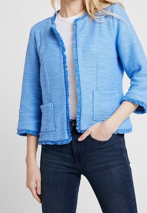 Vrouw draagt een lichtblauwe gestructureerde open-front jas met franjes, een wit overhemd en donkerblauwe spijkerbroek, hand in de zak.