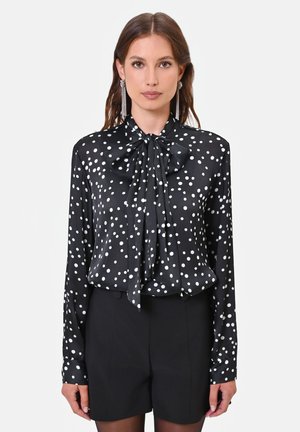 Donna che indossa una camicetta nera a maniche lunghe con pois bianchi e un grande fiocco al collo, abbinata a pantaloncini neri.