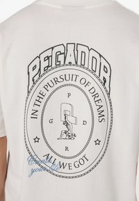 White cotton T-shirt featuring black printed graphic with circular design, "PEGADOR," and "IN THE PURSUIT OF DREAMS," with small blue script detail.