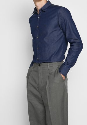 Man draagt een nauwsluitend donkerblauw overhemd met knopen, ingestopt in hoge grijze geplooide broek met handen in de zakken.