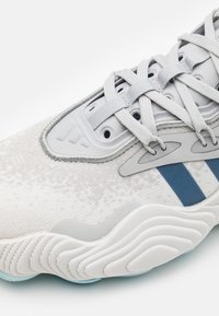 Light grey sneaker with textured fabric upper, blue stripes, and rubber outsole featuring wavy grooves. Detailed lace system and padded collar.