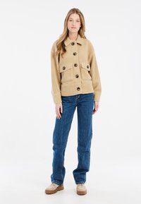 Beige fleece jakke med store frontlommer, knaplukning og krave. Kombineret med blå denimjeans og beige sko.