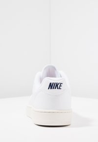 Zapatilla Nike blanca, con parte superior de cuero suave, forma redondeada, costuras visibles y un logo azul marino en el talón, junto con una suela blanca texturizada.