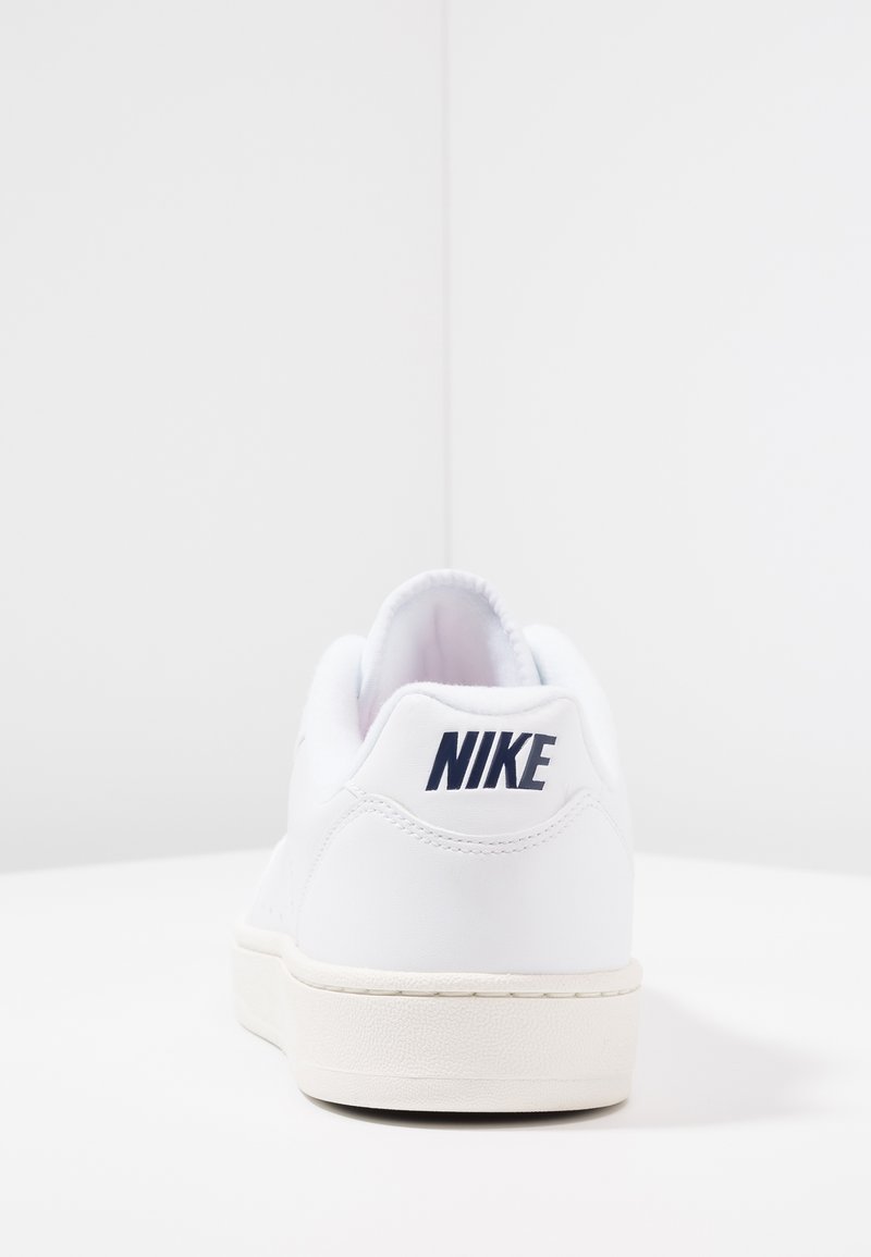 Baskets Nike blanches, tige en cuir lisse, forme arrondie, coutures visibles, et logo bleu marine à l'arrière avec une semelle blanche texturée.