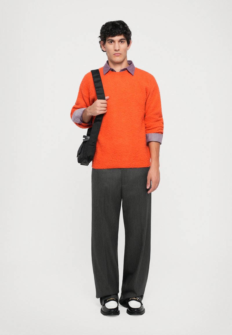 Jeune homme portant un pull orange sur une chemise violette, un pantalon large gris, des mocassins noir et blanc, et portant un sac à bandoulière noir.
