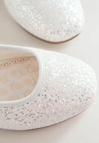 Scarpe basse bianche decorate con glitter iridescenti sparsi. Superficie testurizzata con una fodera interna liscia che presenta un motivo a foglia.