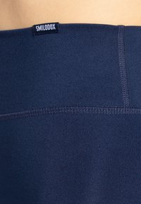 Tmavě modré legíny vyrobené z hladké, pružné tkaniny, s brandovanou etikou na pásku a plochým švem pro zvýšení trvanlivosti.