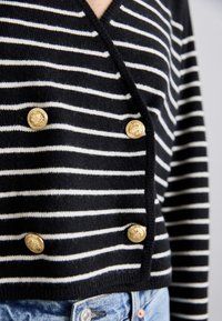 Černobíle pruhovaný pletený cardigan s V-výstřihem, zdobený zlatými knoflíky a zkráceným střihem. Měkká textura a pohodlný střih.