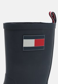 Botte de pluie en caoutchouc noire avec un patch logo rectangulaire en bleu marine, rouge et blanc. Inclut une languette de traction rayée rouge et blanche.