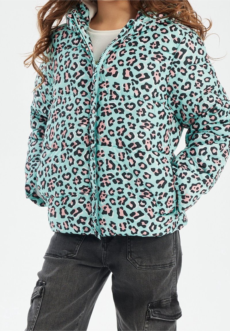 Kind trägt eine türkisfarbene Steppjacke mit pinkem und schwarzem Leopardenmuster, Hände in den Taschen, kombiniert mit schwarzen Cargo-Hosen.