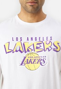 Bijela majica kratkih rukava s odvažnim, šarenim natpisom "Los Angeles Lakers" u ljubičastoj i žutoj boji, s grafikom košarkaške lopte na dnu.