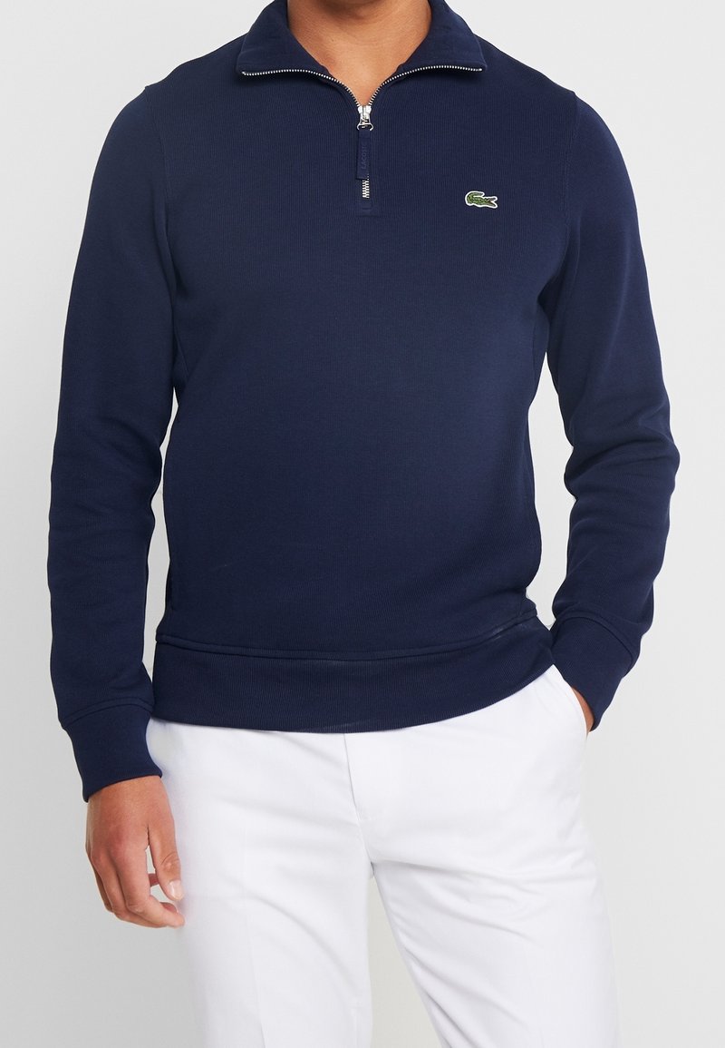 Homme portant un pull bleu marine à demi-fermeture éclair avec un petit logo vert sur la poitrine et un pantalon blanc, les mains dans les poches.