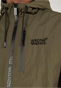 Olivgrüne Jacke aus einem strukturierten, leichten Stoff. Mit einem vorderen Reißverschluss, gesticktem Logo "WRONG FRIENDS" und verstellbarem Kapuzenzug.