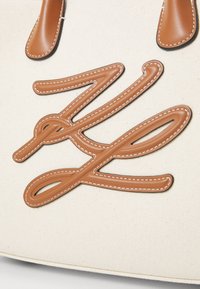 Lærredstoftaske med et stort, syet logo design i brun læder. Tasken har læderhåndtag og en cremefarvet lærredskrop.